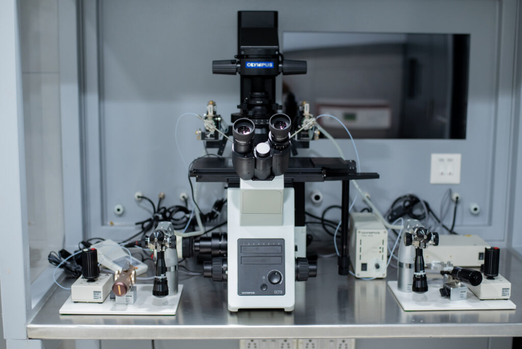 Close-up view of an advanced Olympus microscope setup inside a sterile lab environment. The equipment includes micromanipulators and microinjectors used in assisted reproductive technologies like IVF and ICSI, neatly arranged on a stainless steel workbench.