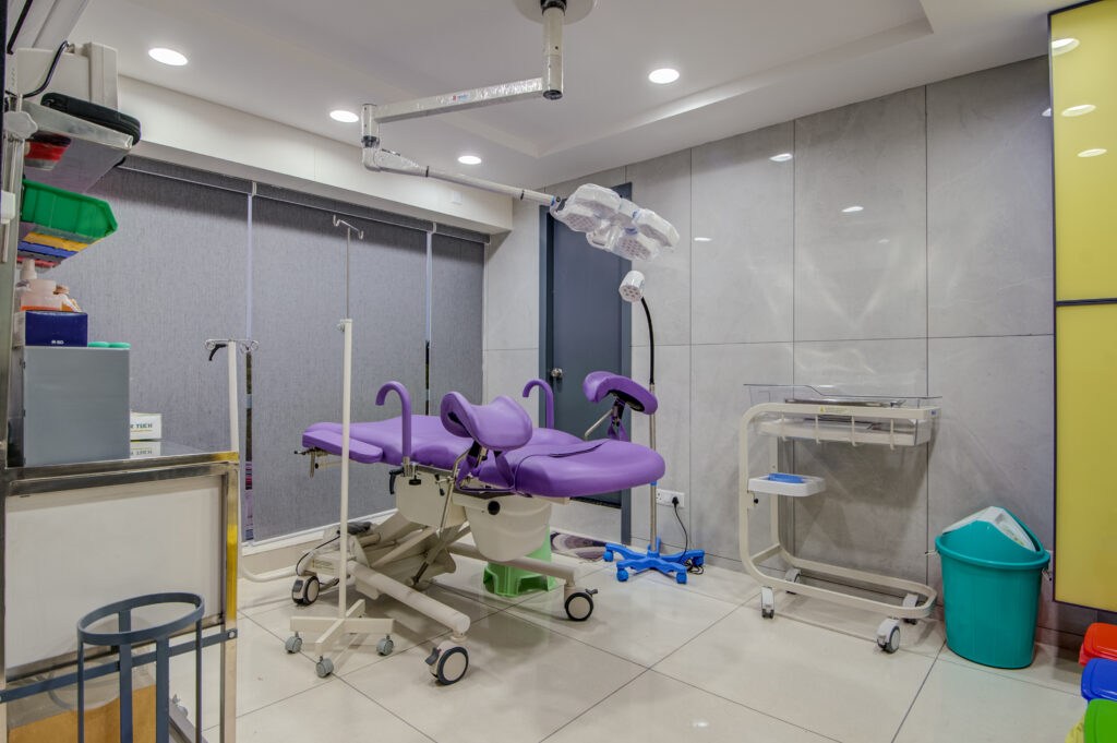 A modern gynecology or fertility treatment room featuring a purple adjustable examination chair with stirrups, ceiling-mounted examination lights, and medical equipment like IV stands, baby warmer, and storage trays. The room has a clean, sterile, and well-organized environment.